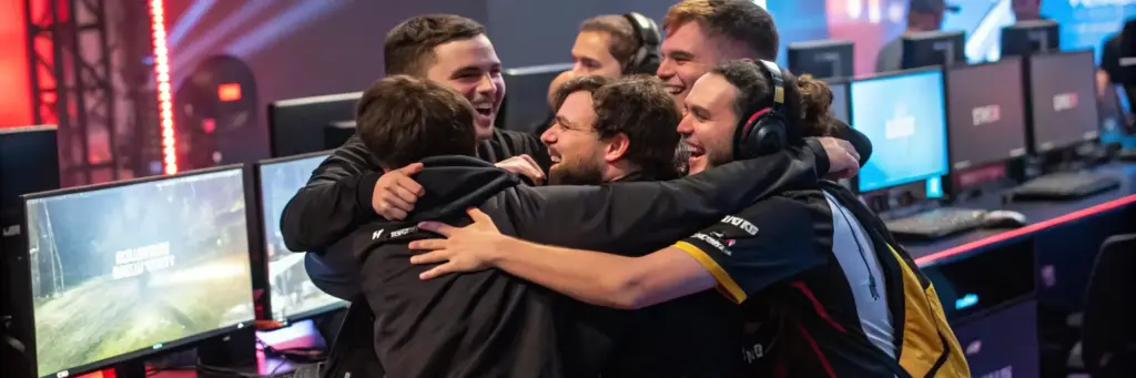 Een groep van zes esports-spelers in teamshirts en met headsets viert het met een groepsomhelzing voor hun computers, waarbij ze hun enthousiasme en teamgeest laten zien