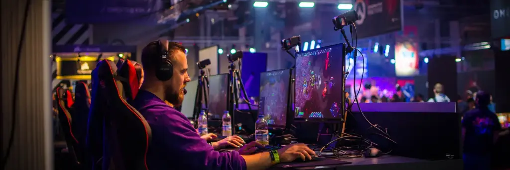 Een geconcentreerde gamer met een koptelefoon zit achter een computer in een drukke, sfeervol verlichte esports-arena. Kleurrijke lampjes en spandoeken zorgen voor een levendige sfeer